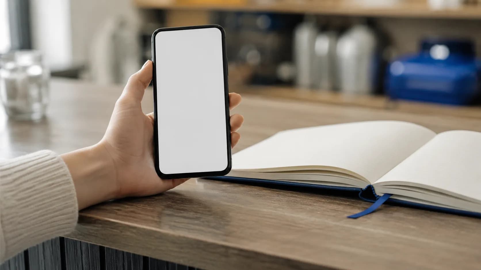 Une main tenant un téléphone à l'écran vide, posée sur un comptoir à côté d'un carnet ouvert.