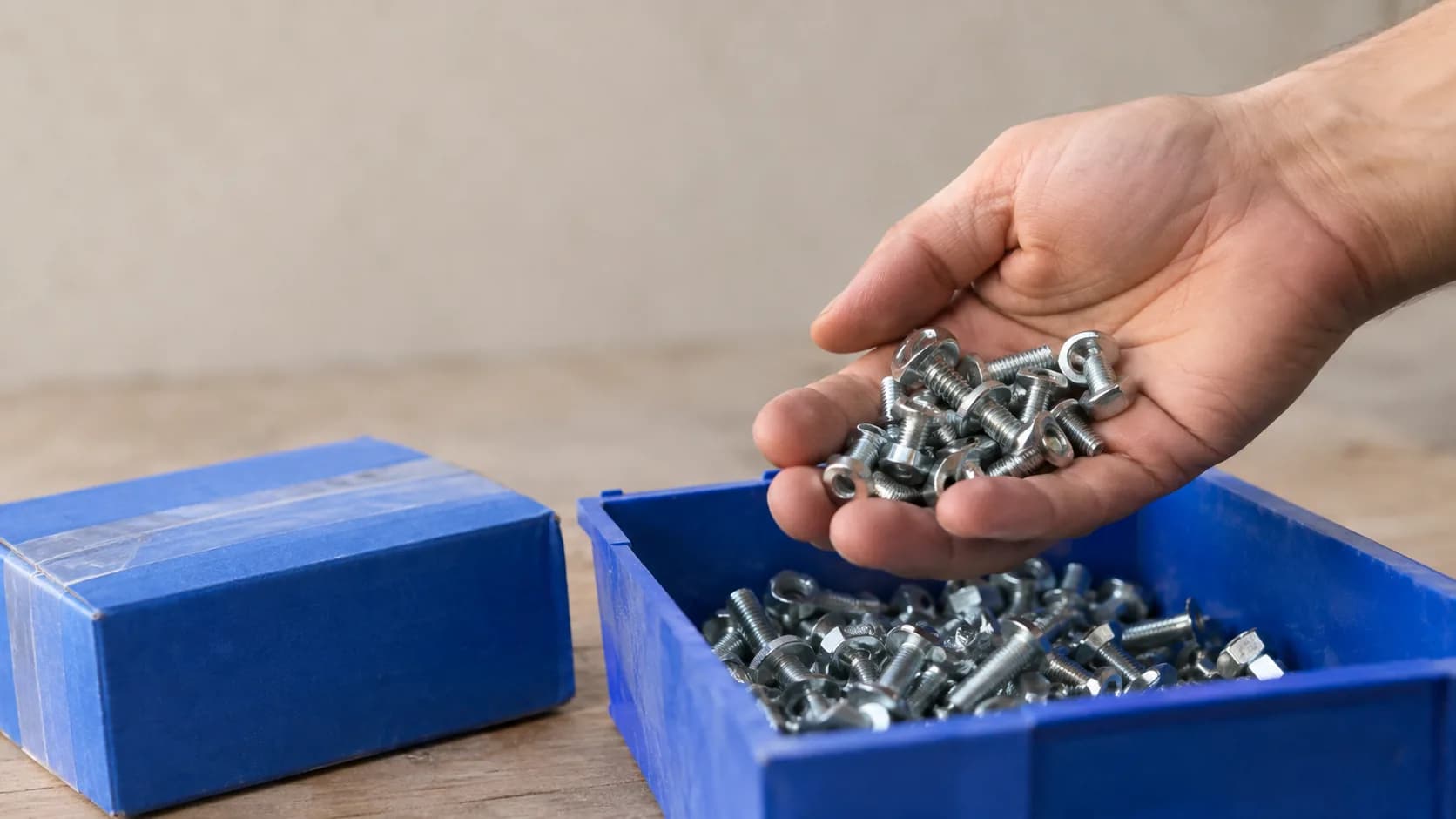 Une main puisant une poignée de petites pièces métalliques dans un bac ouvert, une boîte scellée du même article posée à côté.