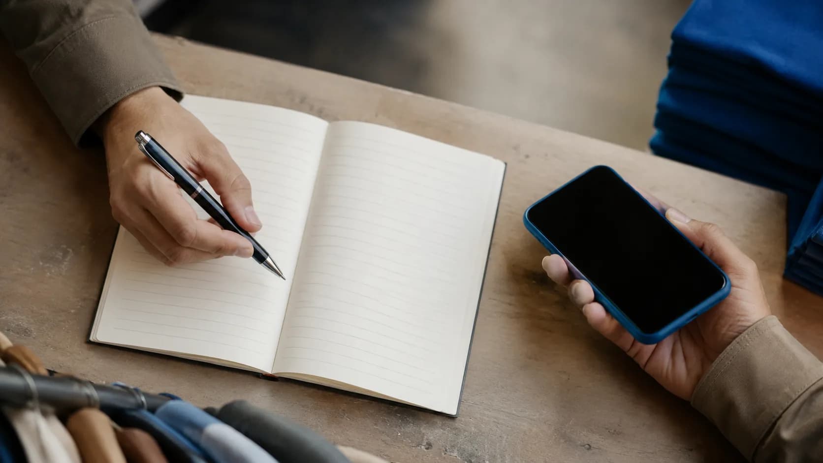 Vue de dessus de deux mains sur un comptoir : l'une tient un stylo au-dessus d'un carnet ligné, l'autre un téléphone.